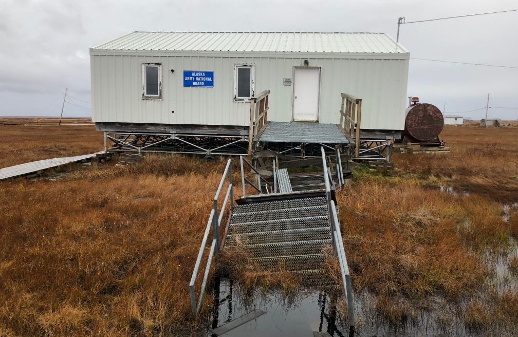 Building in field surrounded by brown grass, staircase sinking into flooded grass