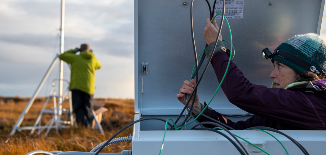 Person standing in a field next to an antenna structure, woman nearby working with wires